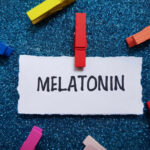 A piece of paper with the word MELATONIN clipped by a red clothespin, surrounded by colorful clothespins on a blue glitter background
