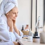 Are Your Skincare Habits Aging You Faster? Woman in a bathrobe with a towel on her head, smiling at her reflection in a mirror