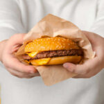 A person holding a cheeseburger wrapped in paper against a yellow background
