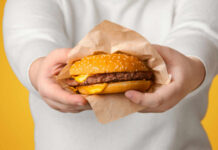 Workplace Diet Trap Exposed A person holding a cheeseburger wrapped in paper against a yellow background