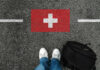Feet wearing white sneakers standing on a road with a painted Swiss flag and a black backpack nearby