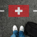 Feet wearing white sneakers standing on a road with a painted Swiss flag and a black backpack nearby