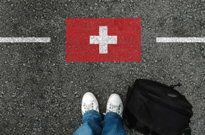 shutterstock_1243055761 Feet wearing white sneakers standing on a road with a painted Swiss flag and a black backpack nearby