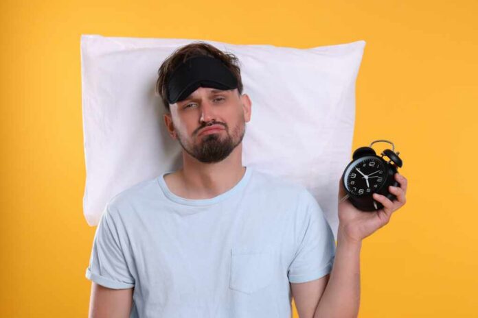 Unhappy,Man,With,Pillow,,Alarm,Clock,And,Sleep,Mask,On Man wearing a sleep mask holding an alarm clock with a frustrated expression