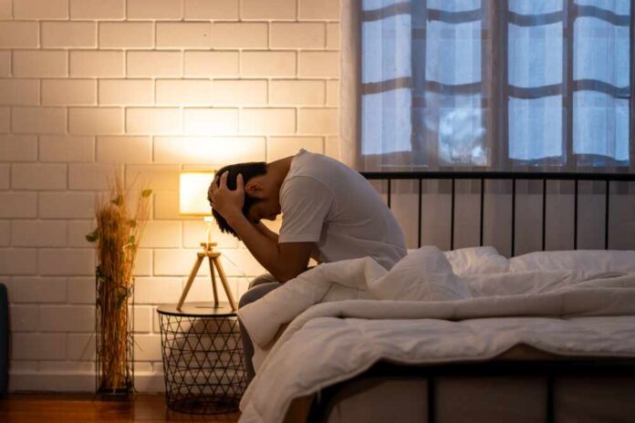 Asian,Frustrated,Insomnia,Man,In,Pajamas,Sit,On,Bed,In A person sitting on a bed with their head in their hands, expressing distress