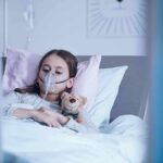 Child in hospital bed with oxygen mask holding teddy bear