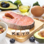 A variety of healthy foods arranged on a table, including salmon, avocado, and nuts, with wooden blocks spelling 'KETO'.