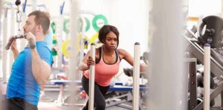 Woman performing sled push in a gym beside a man using a cable machine