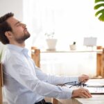 Man relaxing at desk with eyes closed beside laptop