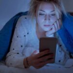 The 20-Minute Insomnia Rule: Get Out of Bed to Sleep Woman under a blanket looking at a smartphone in bed