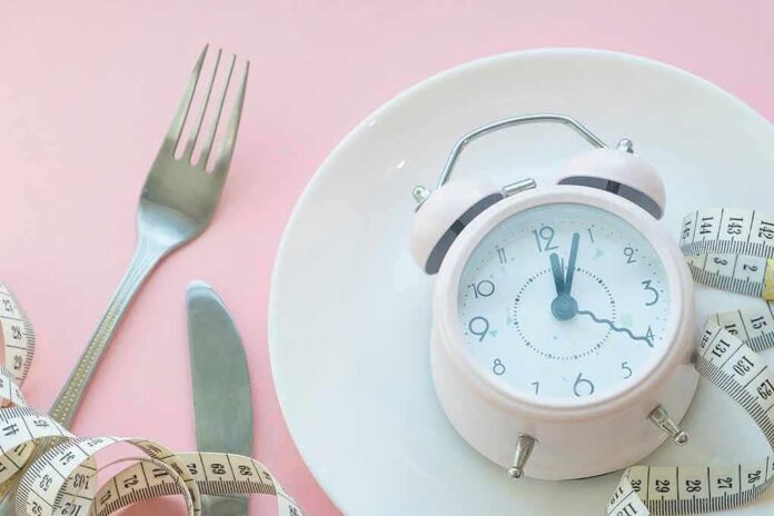shutterstock_1257924982 Pink alarm clock on a white plate with fork, knife, and measuring tape