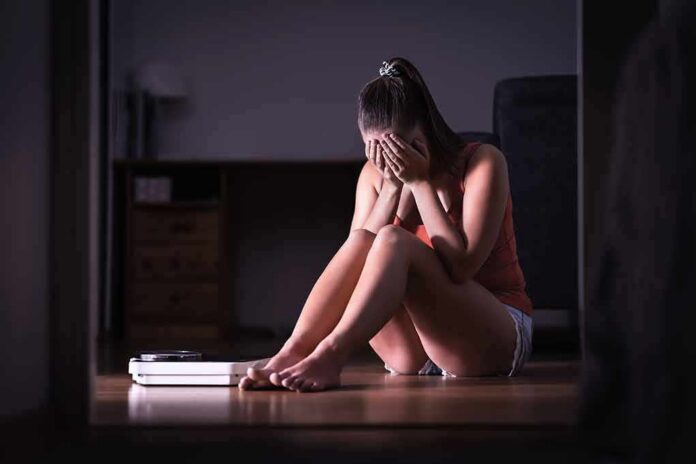 shutterstock_1291372702 Woman sitting on floor covering face beside a bathroom scale