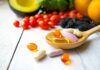 Wooden spoon holding assorted vitamin pills beside fresh fruits and vegetables