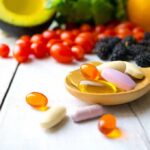 Wooden spoon holding assorted vitamin pills beside fresh fruits and vegetables