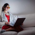 Person wearing orange glasses using a laptop on a white couch