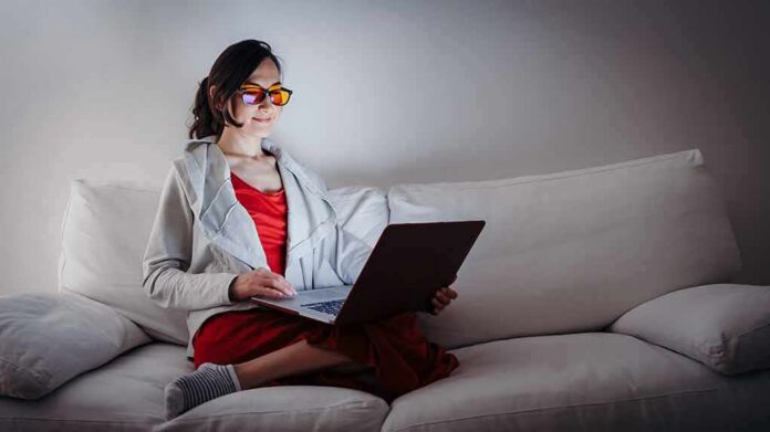 Person wearing orange glasses using a laptop on a white couch