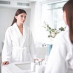 Woman in white bathrobe looking at herself in a bathroom mirror