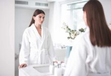 Woman in white bathrobe looking at herself in a bathroom mirror
