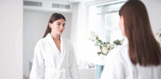 Woman in white bathrobe looking at herself in a bathroom mirror