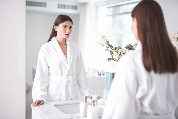 shutterstock_1166393167 Woman in white bathrobe looking at herself in a bathroom mirror
