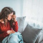 Woman sitting on couch holding pills and rubbing her forehead