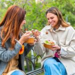 Two women sitting on a park bench eating takeout bowls