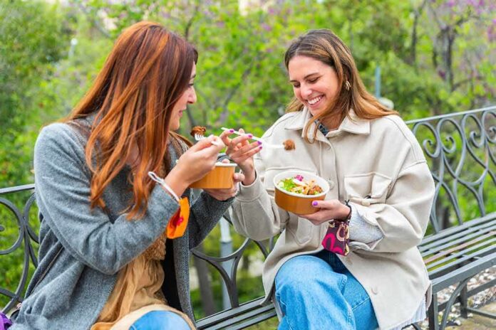 shutterstock_2049883694 Two women sitting on a park bench eating takeout bowls