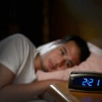 Person in bed looking at a digital alarm clock reading 2:21